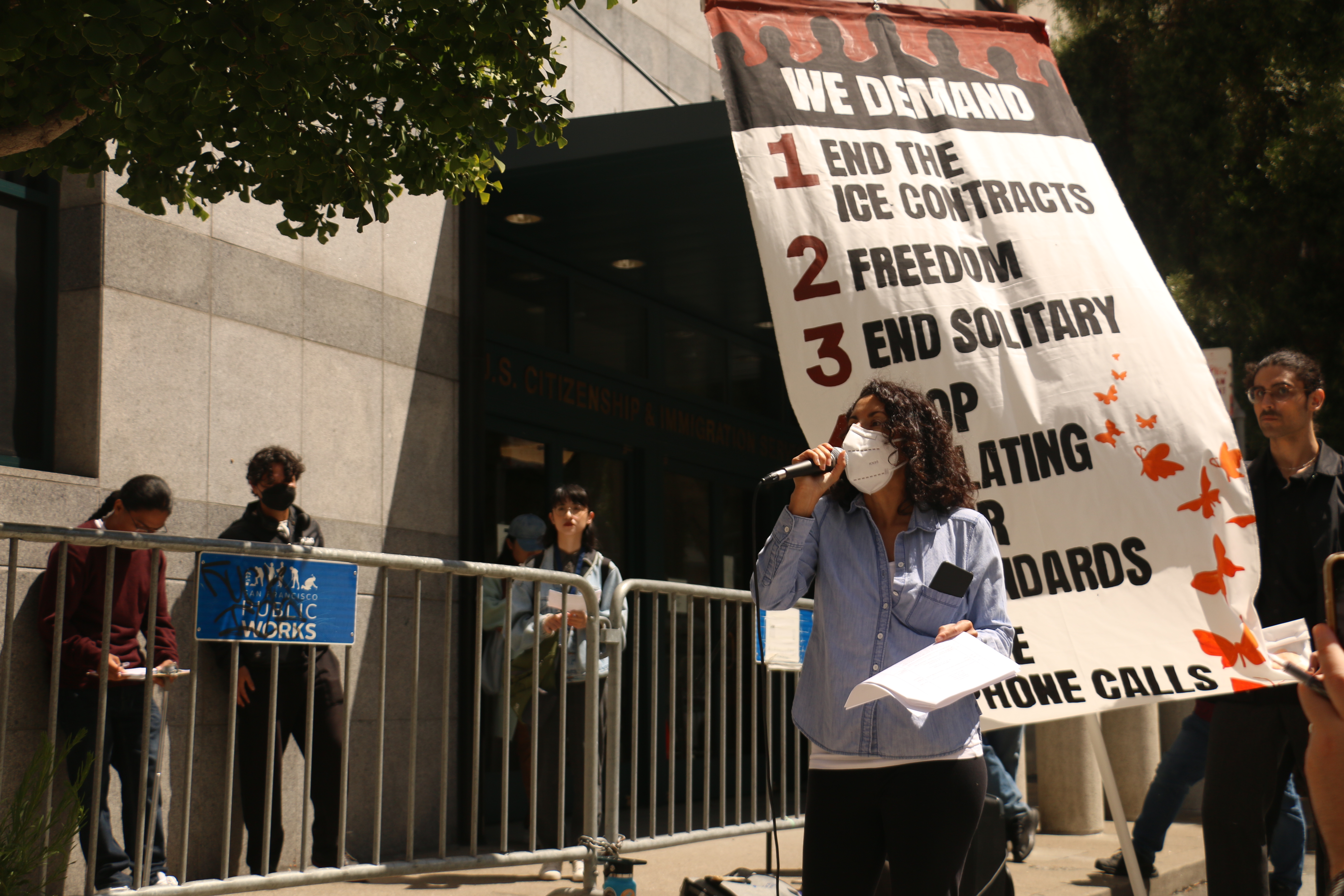 A woman wearing a mask holds papers and looks away from the camera as she speaks into a microphone. A supporter holds a large vertical banner behind her with the following demands painted: "End the ICE Contracts / Freedom / End Solitary / Stop Violating Your Standards / Free Phone Calls"