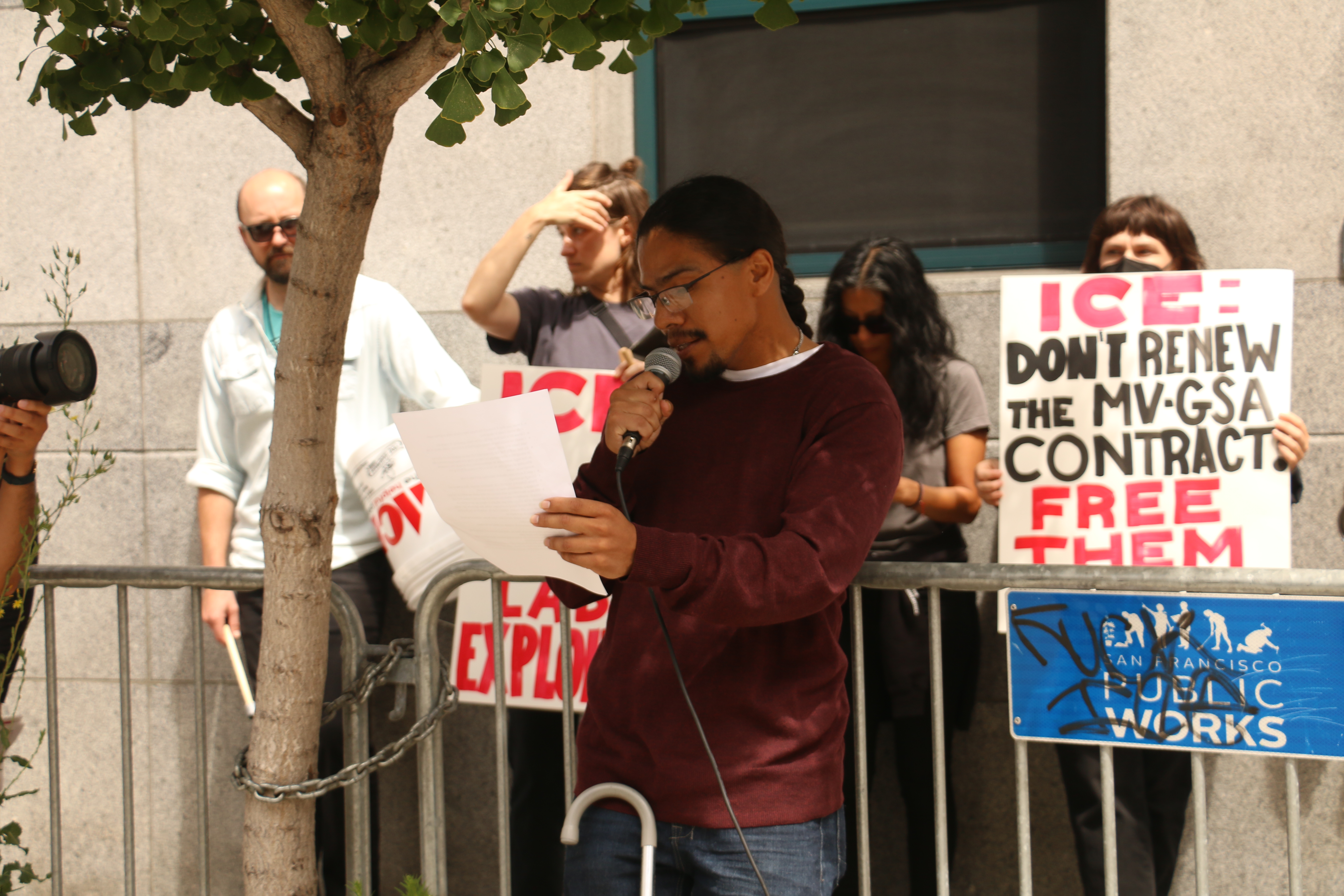 Jose Ruben Hernandez Gomez wears glasses and speaks into a microphone. His mobility device rests on his leg as he reads off of a paper. Behind him are people holding signs. One says, "ICE! Don't renew the MV-GSA Contract. Free them All."