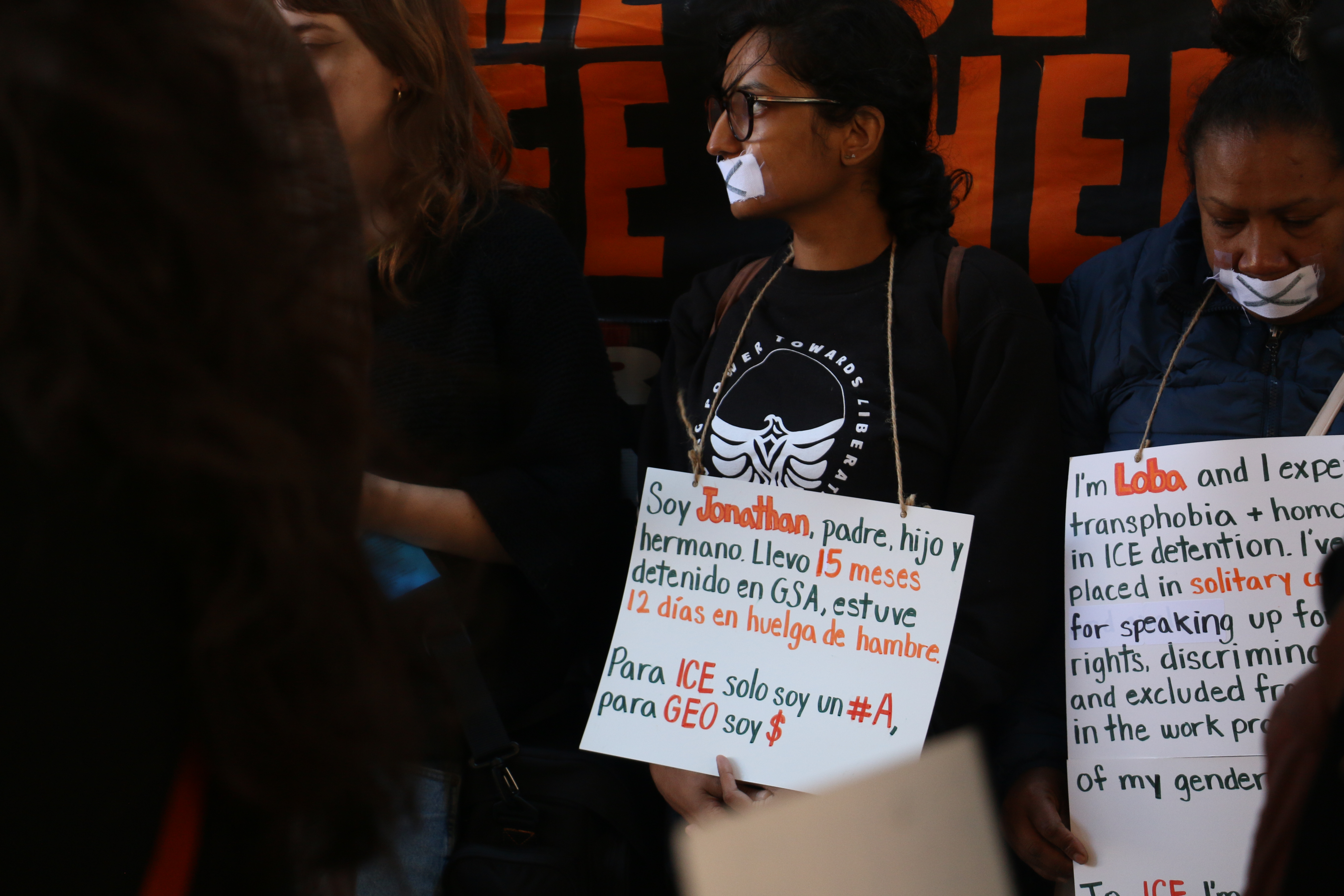 A few supporters stand in front of a large banner. They wear tape over their mouths with "x's" marked over the tape. Signs with the words of people in detention hang around their shoulders. One says, "Soy Jonathan, padre, hijo, y hermano. Llevo 15 meses detenido en GSA, estuve 12 dias en huelga de hambre. Para ICE solo soy un #A, para GEO soy $. / I'm Jonathan, a father, son and brother. I have been detained at GSA for 15 months. I was on hunger strike for 12 days. For ICE I am only an A number, for GEO I am a dollar sign." Another sign begins, "I'm Loba and I experienced transphobia and homophobia in ICE detention..."