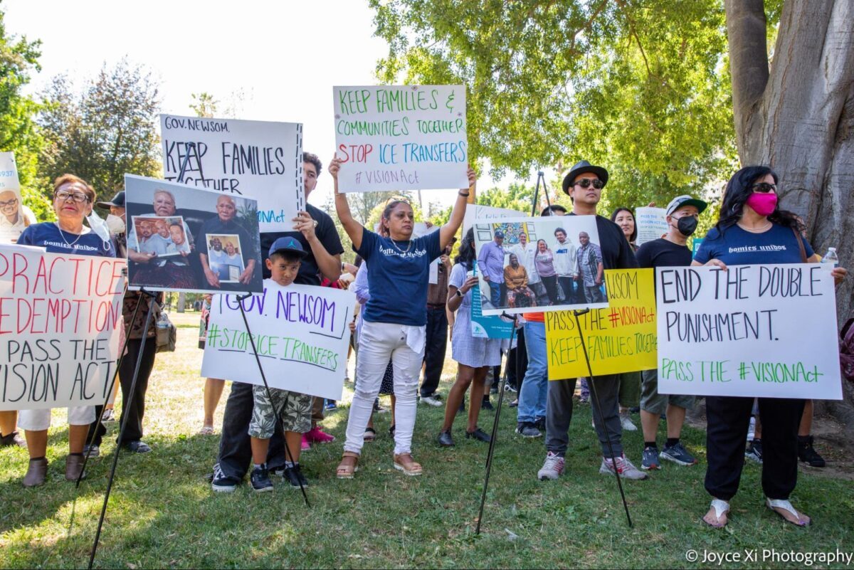 Community members in a park in front of Sacramento's Capitol hold up signs in support of the VISION Act.