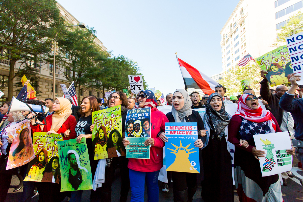 Community members march together at a No Muslim Ban Ever rally, holding loudspeakers and colorful signs that read "refugees welcome" "No Muslim Ban Ever" and more.