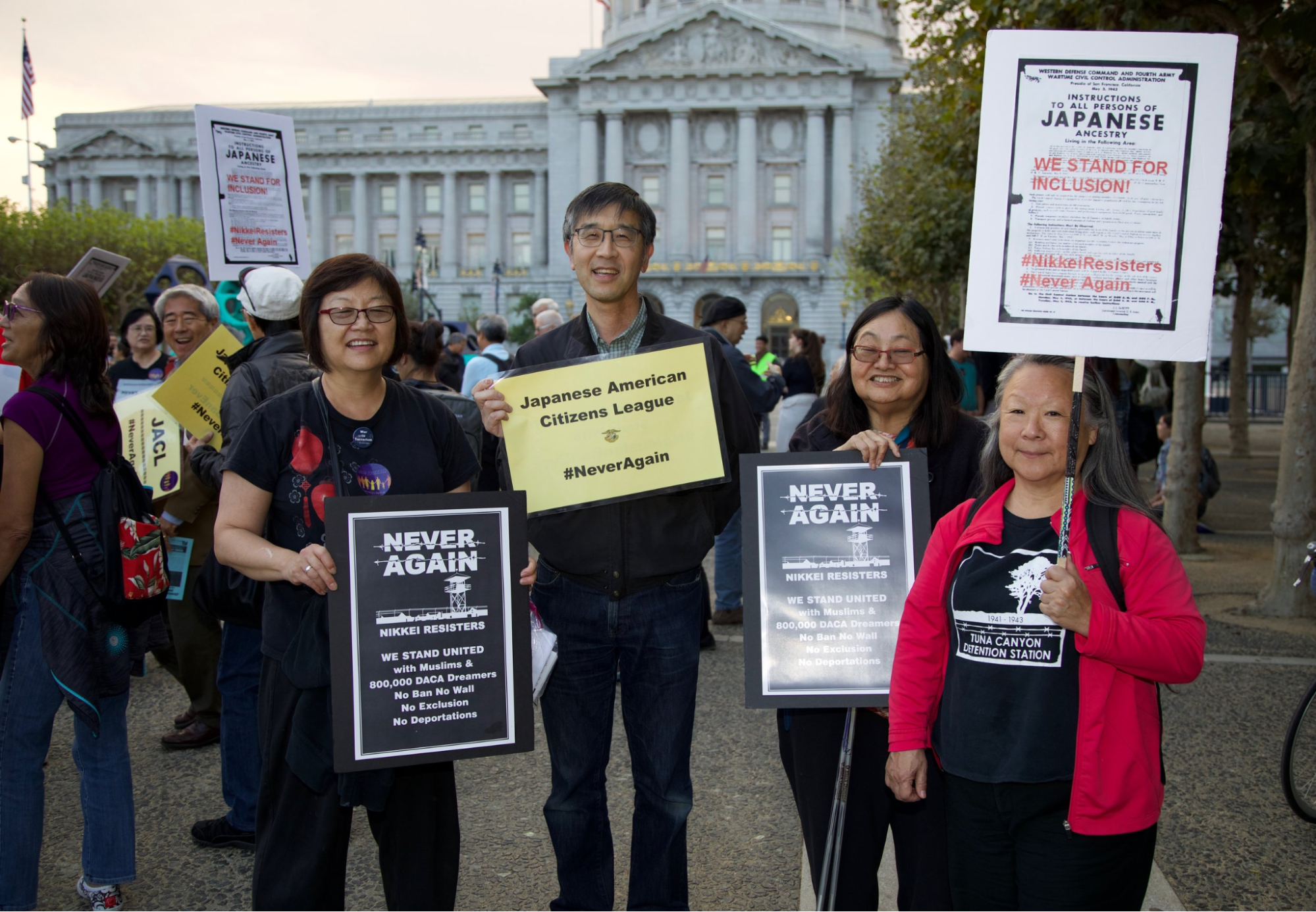Four members of the Japanese American stand in support with the Muslim community at a rally to end the Bans, holding signs that say "Never Again"