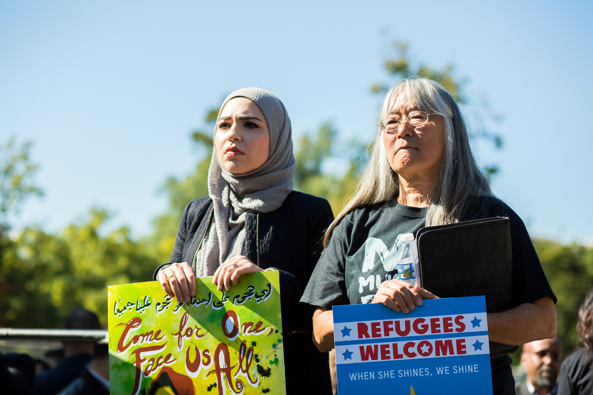 A woman in a gray hijab and a Japanese woman with gray hair and black t-shirt stand together at a No Muslim Ban protest.