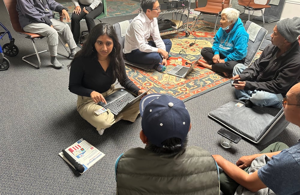 A photo shows several people sitting cross legged on a carpeted floor. Two sit in front of laptops as they speak to different people.