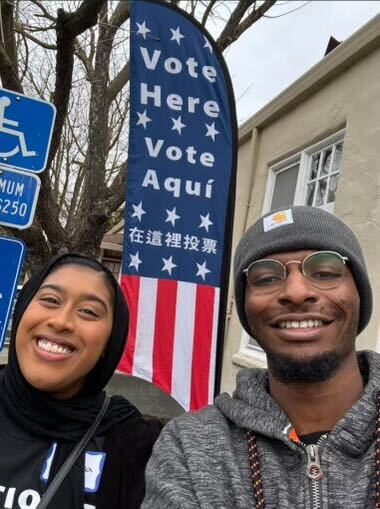 2 poll monitors posing in front of a sign that reads 'Vote Here' and 'Vote Aqui'.