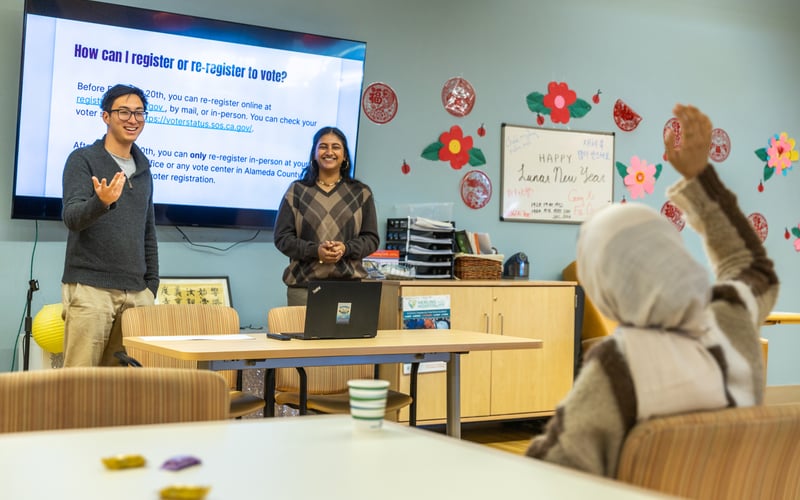 In February 2024, ALC staff held a workshop on voting rights for community members at On Lok in San Jose. In the photo background, two ALC staff members stand on either side of a slide presentation. The slide says "how can I register or re-register to vote?" In the foreground, a community member raises their hand to ask a question.