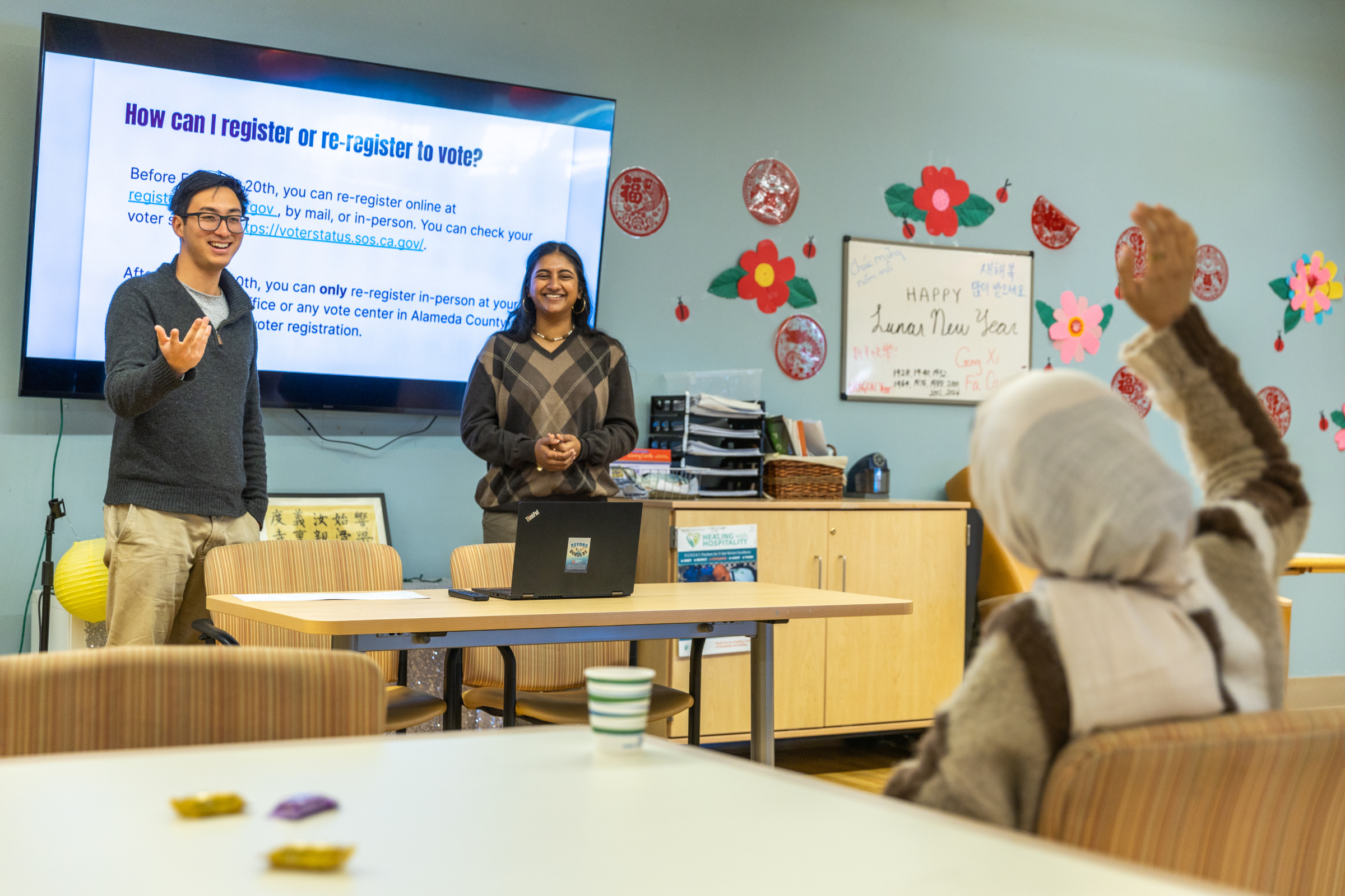 In February 2024, ALC staff held a workshop on voting rights for community members at On Lok in San Jose. In the photo background, two ALC staff members stand on either side of a slide presentation. The slide says "how can I register or re-register to vote?" In the foreground, a community member raises their hand to ask a question.