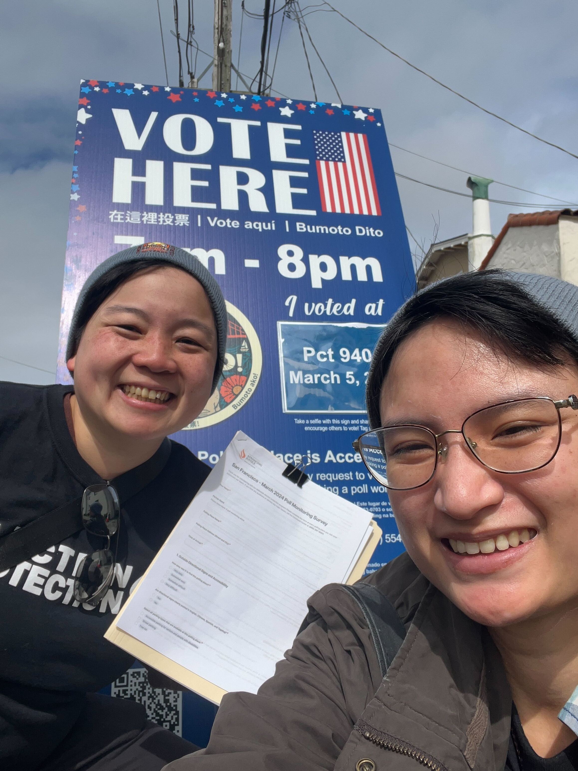 2 poll monitors posing in front of a sign that reads 'Vote Here'.