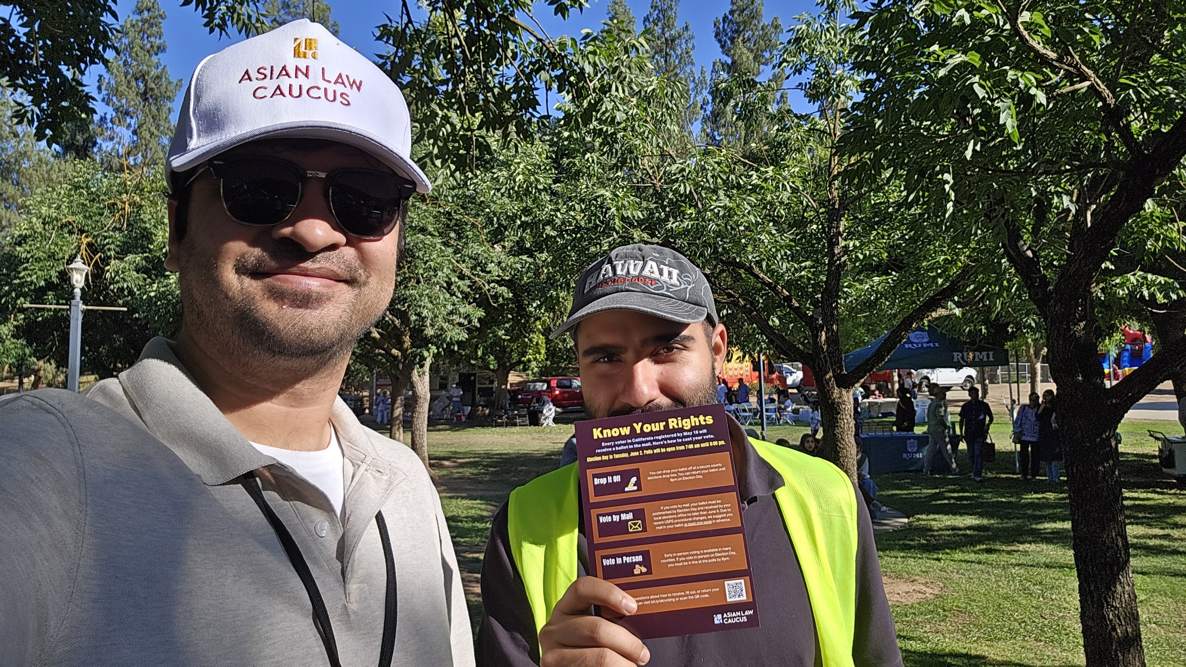 Two men wearing baseball caps pose for a selfie photo. One wears a hat that says Asian Law Caucus with a lanyard and the other holds up a card with Know Your Rights information on it. They stand outside in a grassy area.