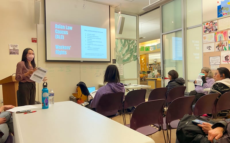 An Asian Law Caucus community advocate stands in front of a room holding papers as she gestures. Behind her is a projector screen hanging from the ceiling with a powerpoint slide that details Asian Law Caucus services around workers' rights. A few rows of people face her, and behind them, several colorful,hand-drawn pictures on 8x11" paper are on display.
