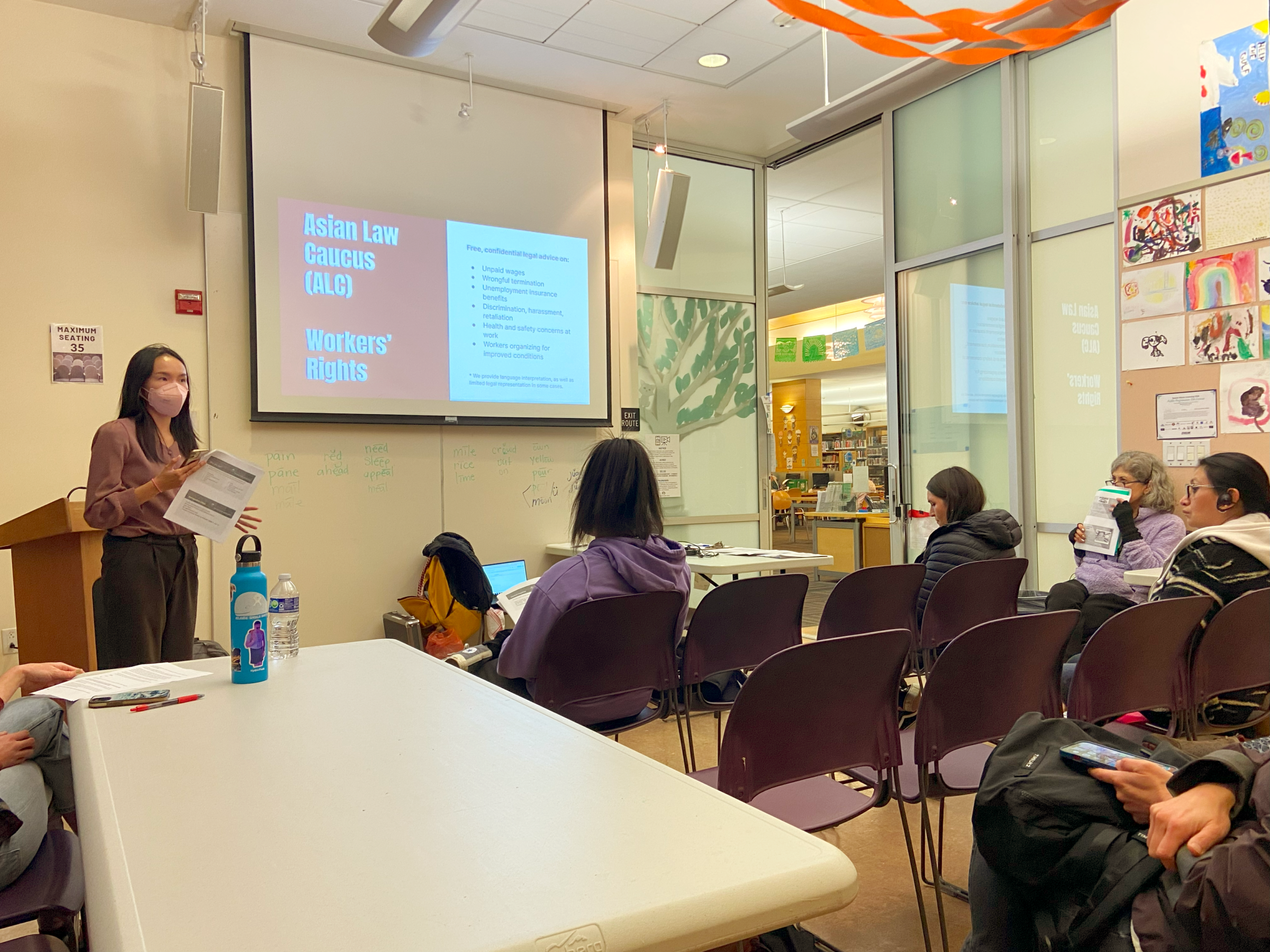 An Asian Law Caucus community advocate stands in front of a room holding papers as she gestures. Behind her is a projector screen hanging from the ceiling with a powerpoint slide that details Asian Law Caucus services around workers' rights. A few rows of people face her, and behind them, several colorful,hand-drawn pictures on 8x11" paper are on display.
