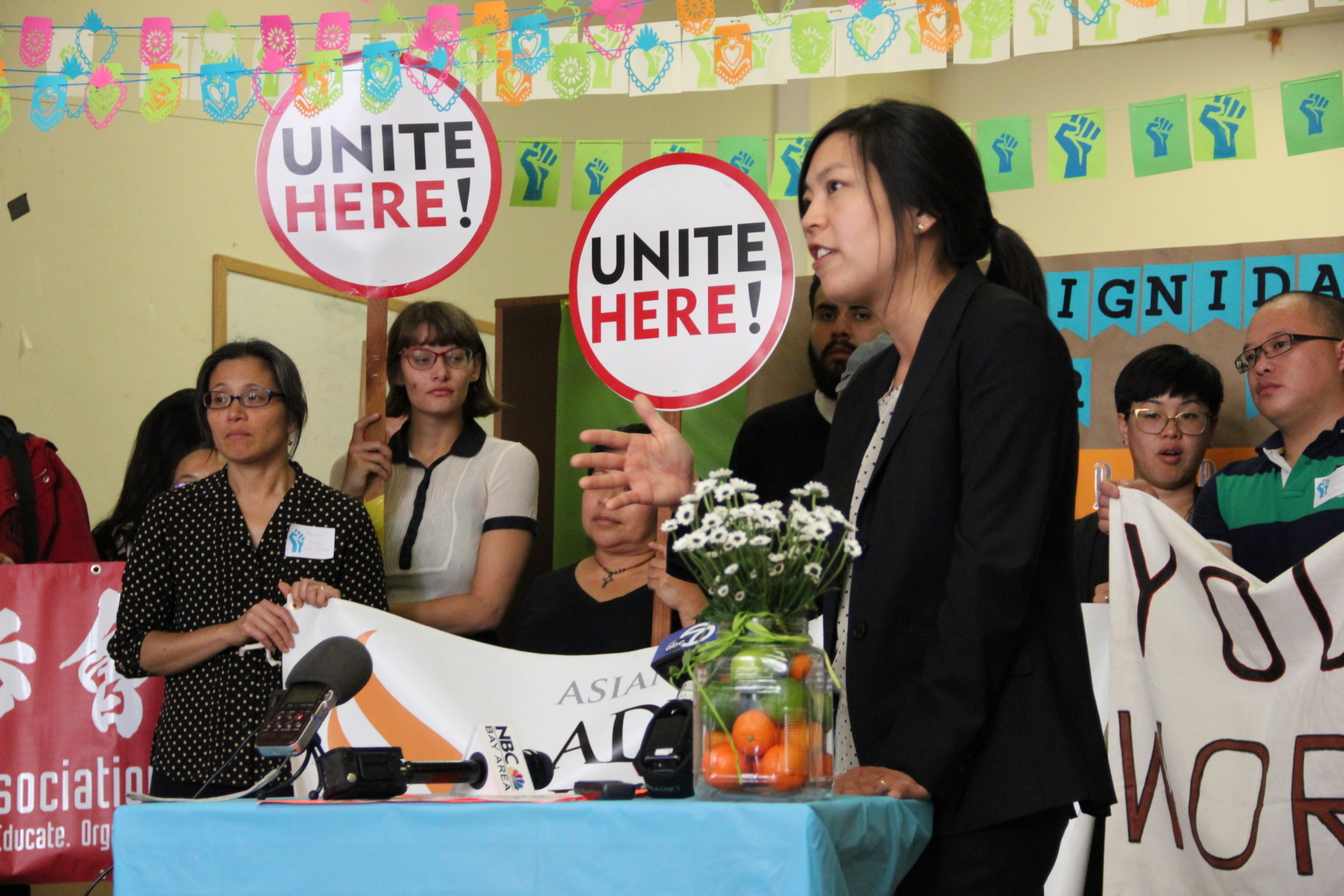 ALC attorney WInnie Kao speaks at a press conference behind a table with microphones and a vase with oranges and flowers. Behind her colorful bunting hangs from the ceilings and people hold banners and signs saying "Unite Here!"