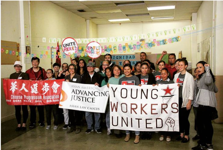 A group of about 20 community members and leaders holding up a fist for victory and holding banners that read "Chinese Progressive Association", "Asian Law Caucus", and "Young Workers United"