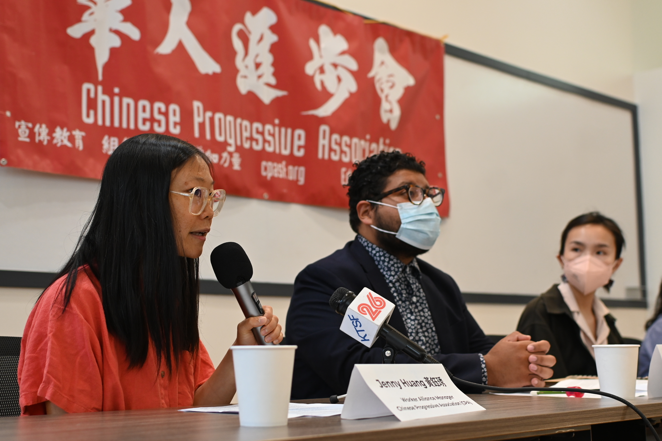 A speaker from Chinese Progressive Association speaks into a microphone in front of a table. Two other sit next to her, facing towards the audience and looking in her direction. In front of the speaker is another microphone from TV outlet KTSF 26. Behind the three people sitting at the table is a large banner that says 'Chinese Progressive Association' in Chinese and English.
