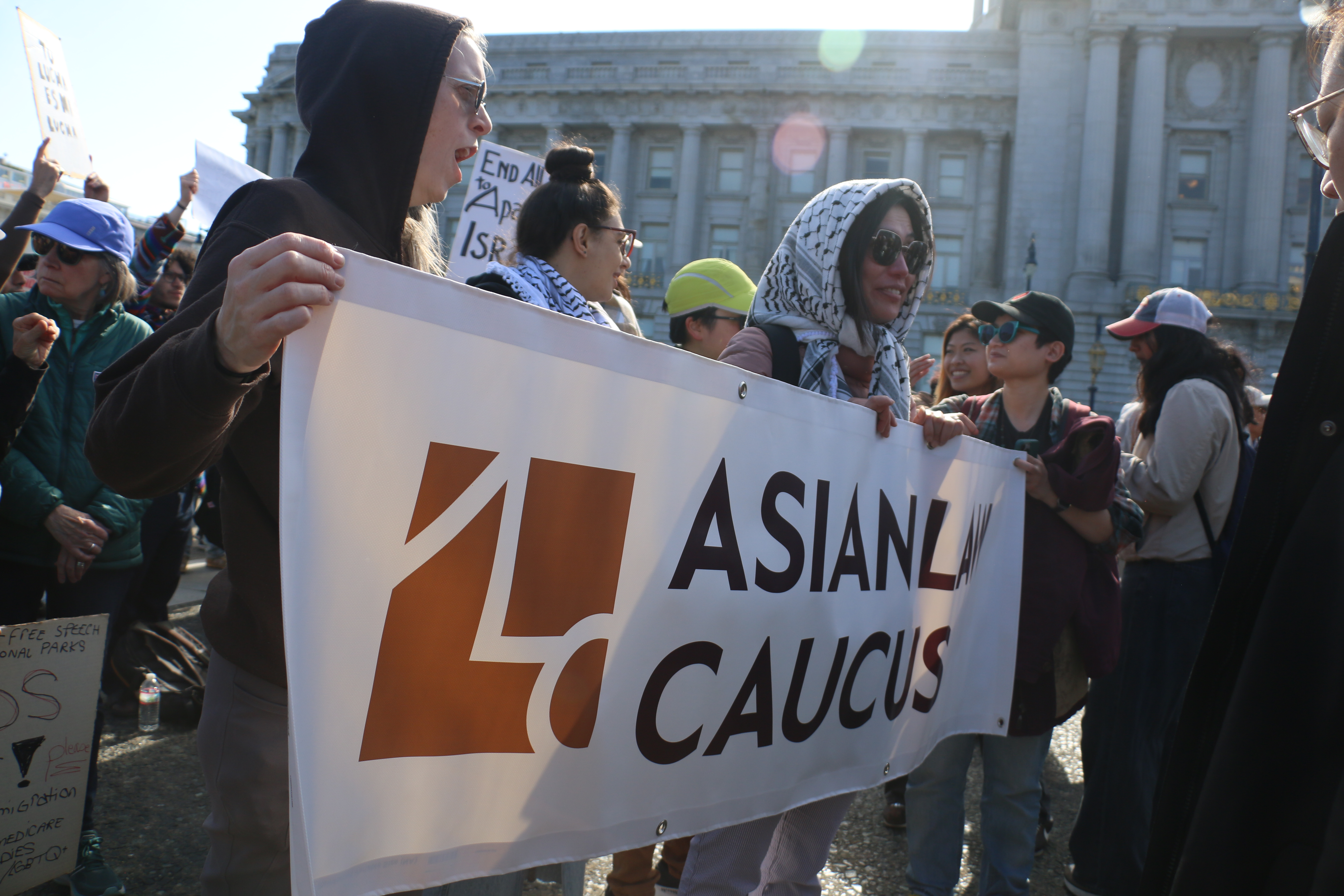 Several people hold up a vinyl banner with the Asian Law Caucus logo in front of San Francisco City Hall. There are other people holding signs in the background.