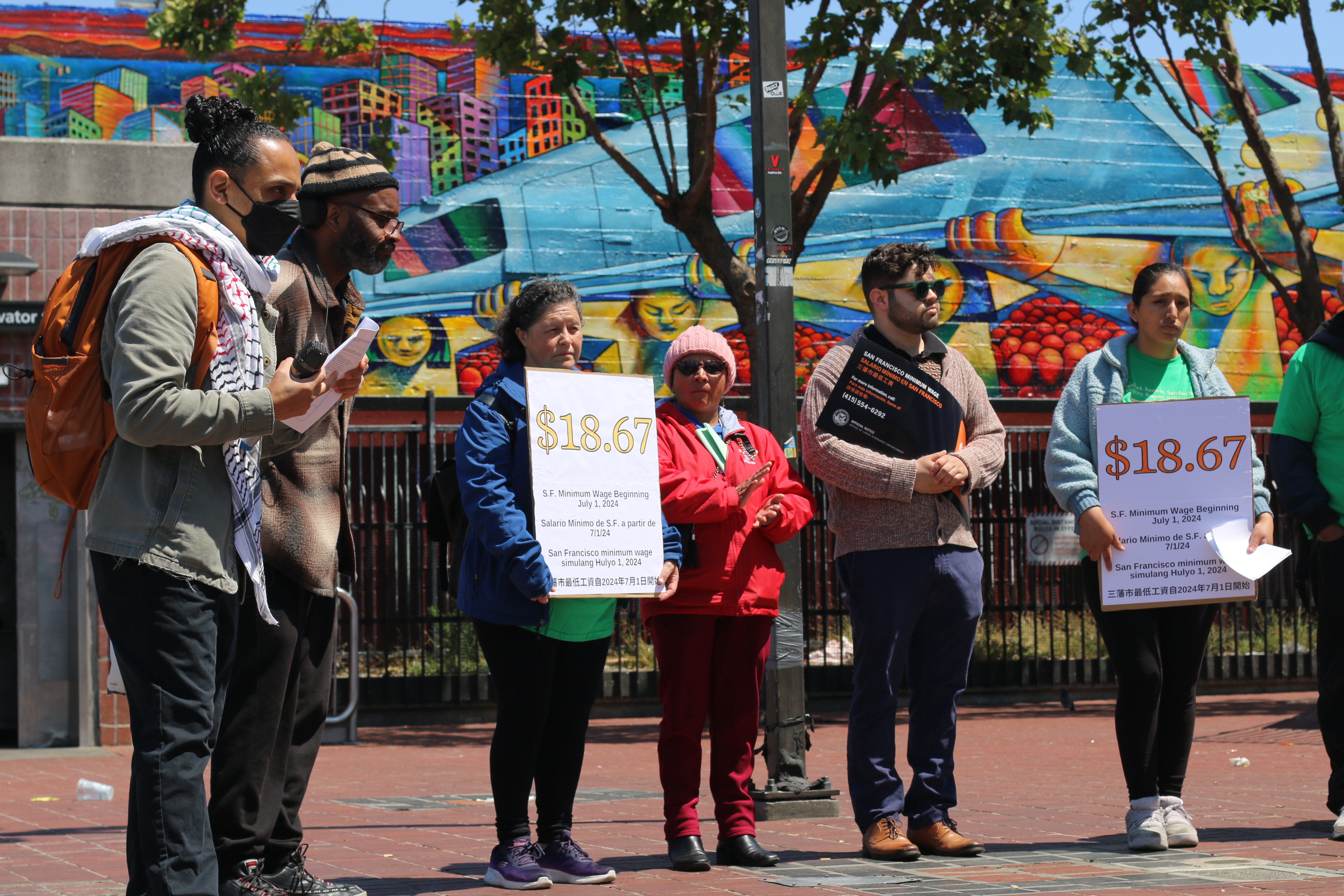 Several people stand together, a large mural of fruits and vegetable and a city landscape behind them. They wear jackets and many hold signs with the San Francisco $18.67 minimum wage printed on them. They look on as someone holding printouts speaks with a microphone in their hand.