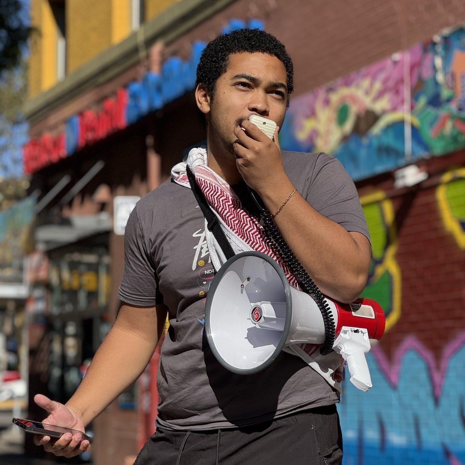 Sabir stands and gestures as he speaks into a megaphone. He wears a keffiyeh across is torso. In the background is a storefront with a colorful mural and graffiti