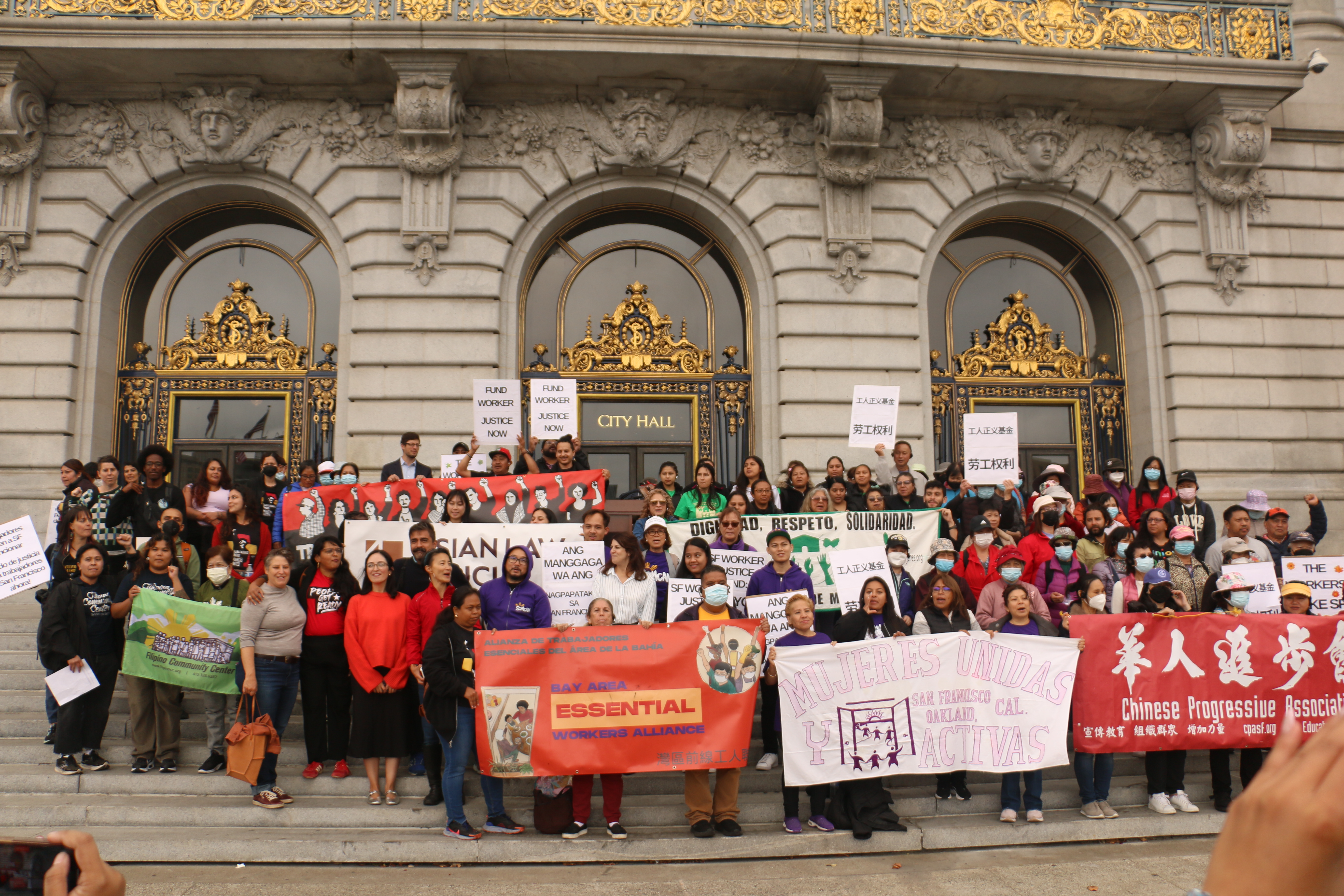 A large crowd gathers on the steps outside San Francisco City Hall, a neoclassical building of stone with gilded metal decorating the entry way and balcony. Several groups of people hold banners of organizations they're part of : Chinese Progressive Association, Bay Area Essential Worker Association, Mujeres Unidas y Activas, Trabajadores Unidos Workers United, Asian Law Caucus, Filipino Community Center, and more. Several hold signs in different languaes that say "Fund Worker Justice Now" and "The Workers Make SF Run."