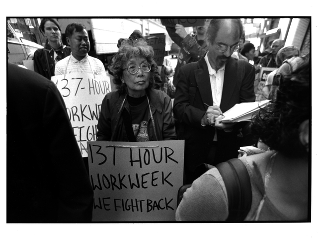 Black and white image shows Yuri at a labor protest holding a sign that reads "131 hour workweek, we fight back".