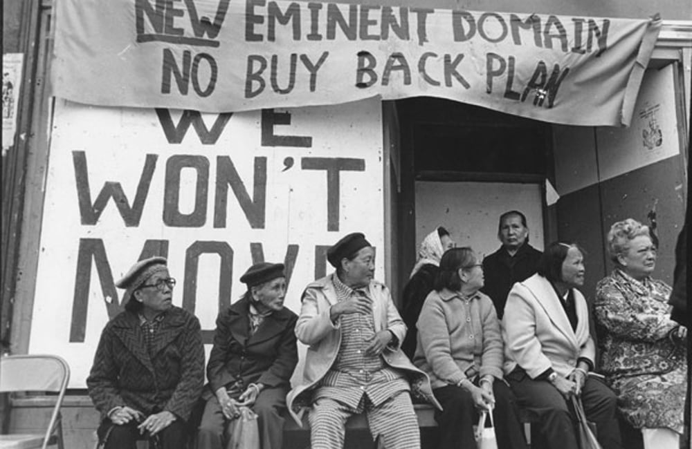 Filipino and Chinese tenants sit outside the I Hotel in San Francisco. Behind them, they are signs that read "New Eminent Domain. No Buy Back Plan." and "We Won't Move."