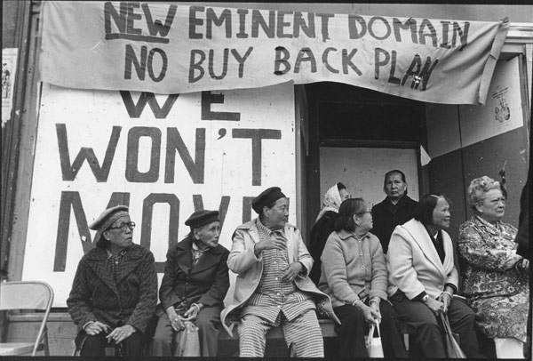 Filipino and Chinese tenants sit outside the I Hotel in San Francisco. Behind them, they are signs that read "New Eminent Domain. No Buy Back Plan." and "We Won't Move."