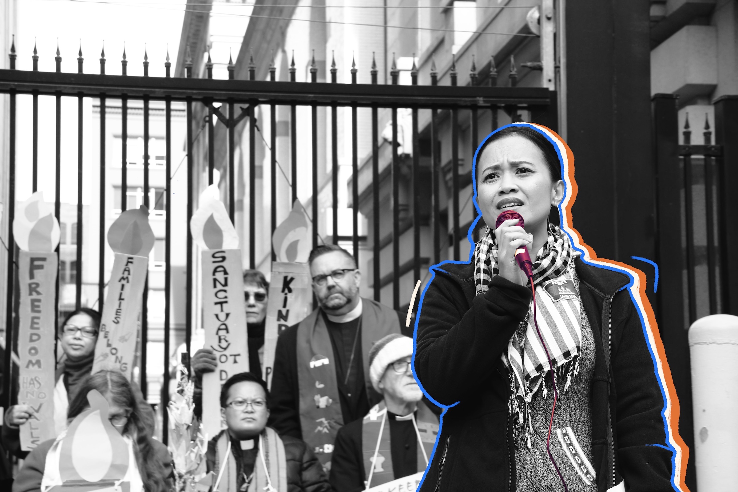 Woman wearing a scarf and black jacket holds a microphone at a rally. A group of community members and faith leaders holding signs stand behind her.