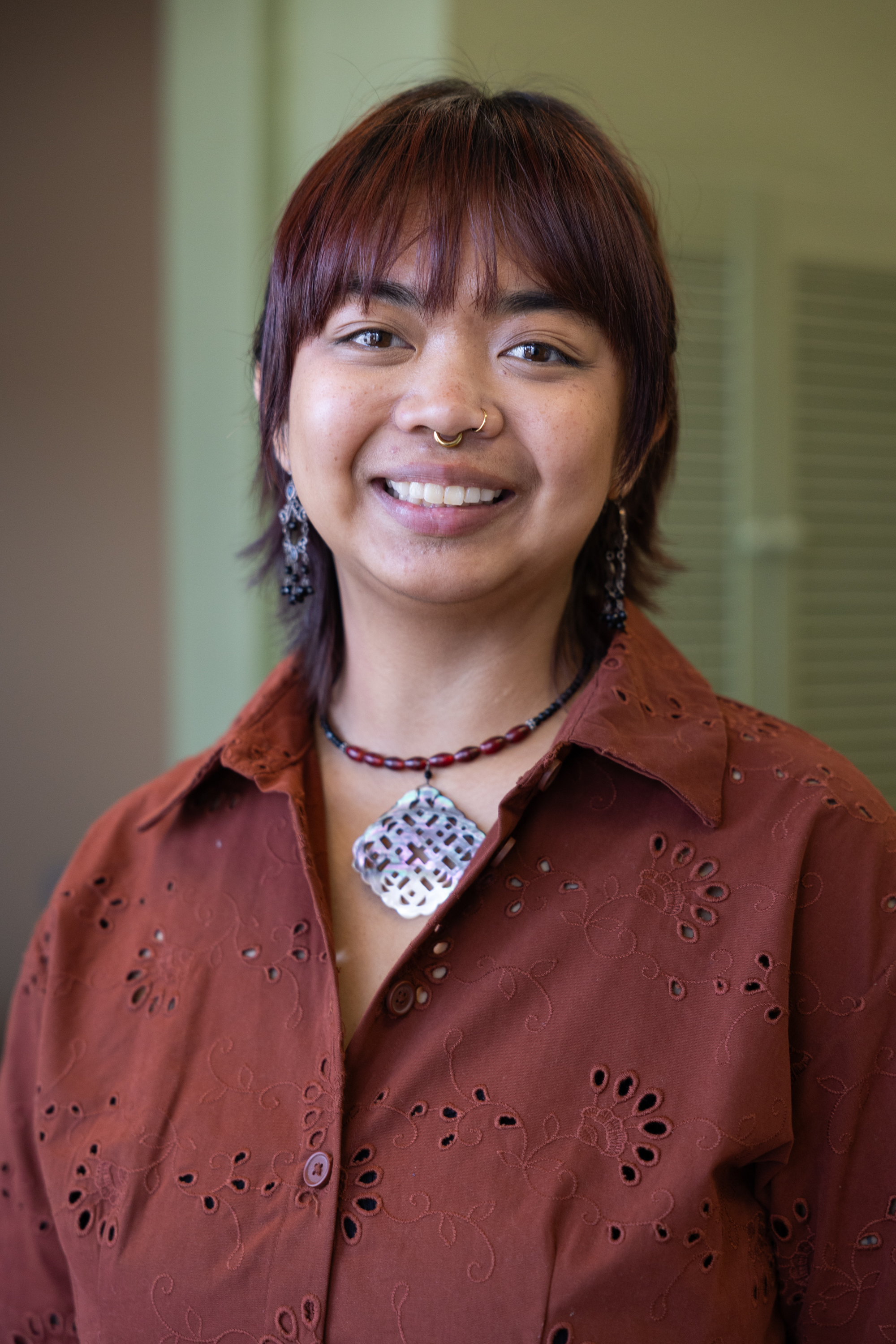 Photo: A person wearing a button-up shirt and necklace smiles at the camera.