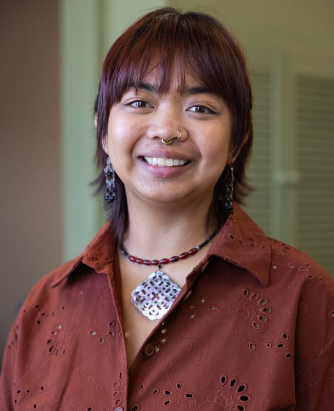 Photo: A person wearing a button-up shirt and necklace smiles at the camera.