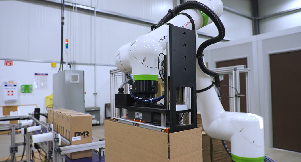 A white and green collaborative robot arm equipped with a custom black end-of-arm tool erects a cardboard box in a warehouse, with a conveyor of sealed cases labeled "RMR" in the background.