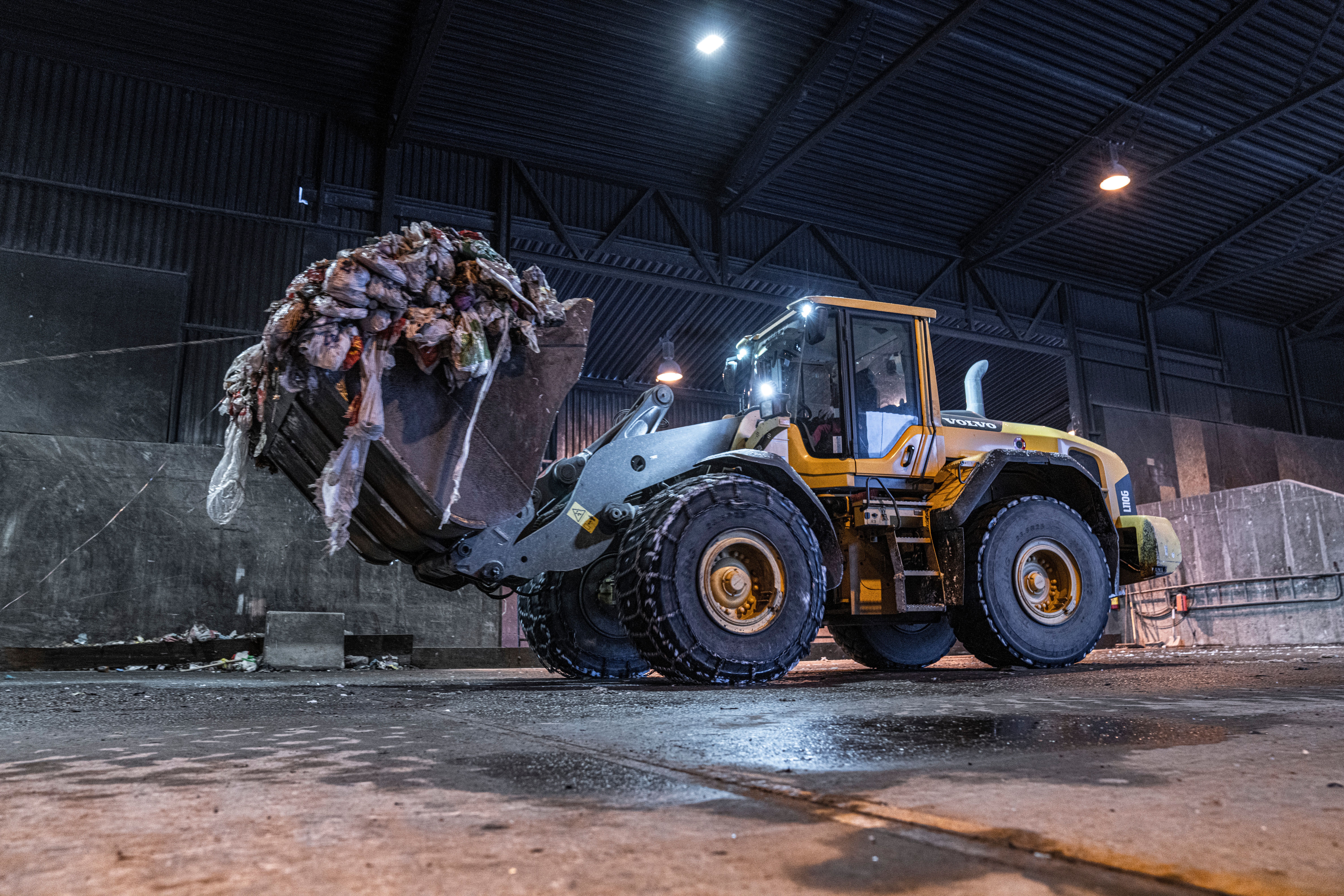 Hjullastar køyrer med frontskuffe full i avfall inne i ein sorteringshall.