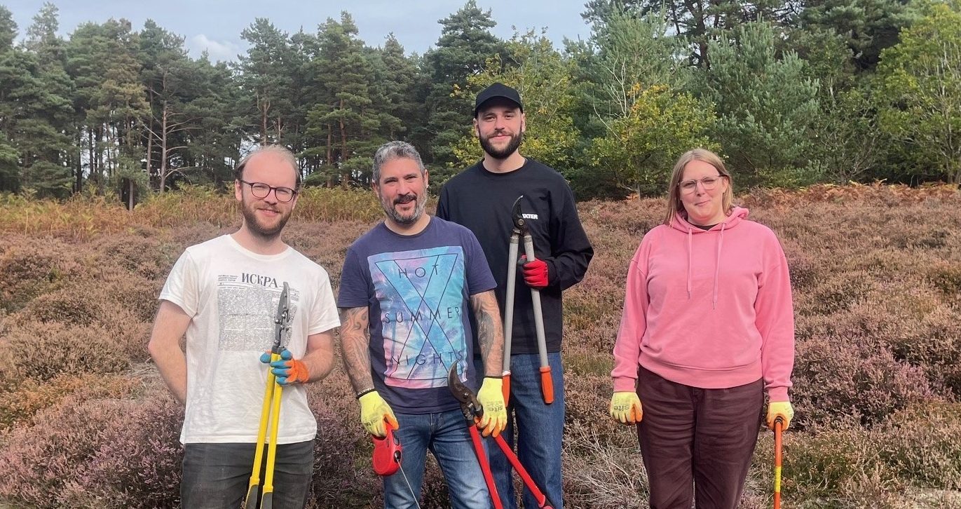 Four people standing in heathland holding long-handled tools, wearing casual clothing and gloves, with pine trees in background.