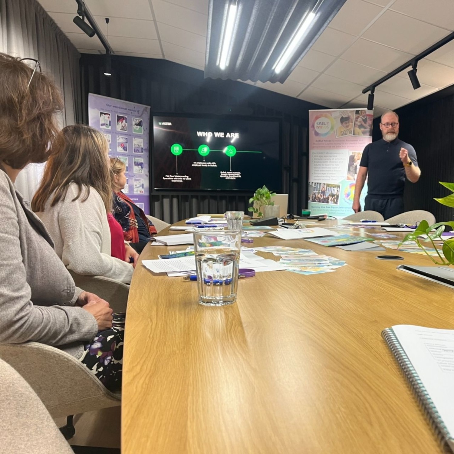 Bearded man in black t-shirt presenting to group seated around wooden table in meeting room with green wall display and papers.