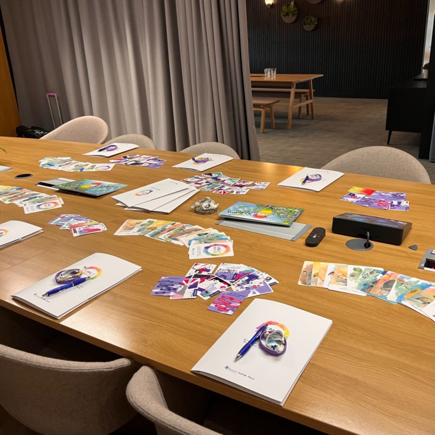 Large wooden table with scattered colourful papers, notebooks, and crafting materials. White chairs around table. Grey curtains and dining area visible in background.