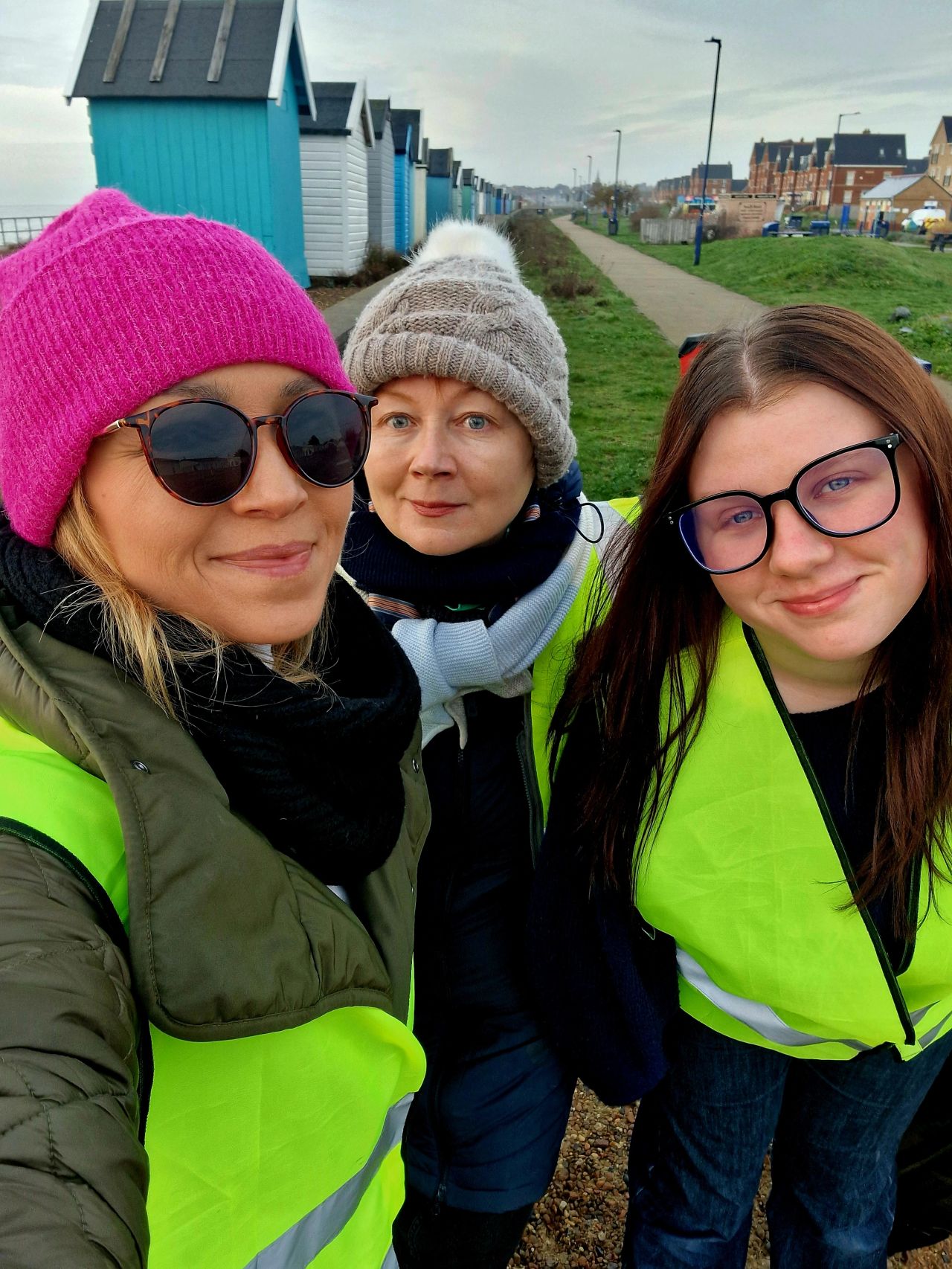 Three women in hi-vis jackets and winter hats taking selfie on coastal path with colourful beach huts and houses in background.