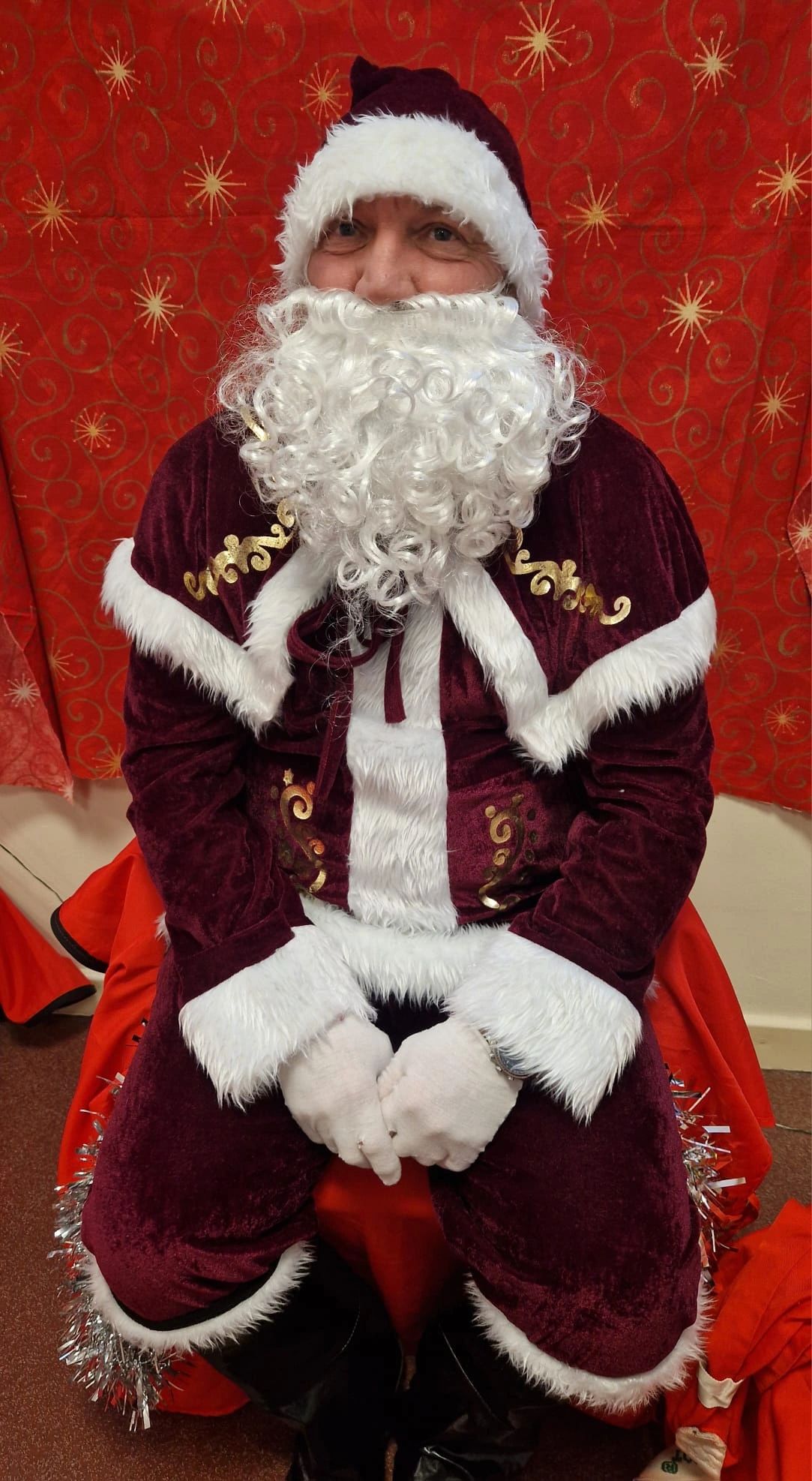Person in Father Christmas costume with burgundy velvet suit, white fur trim, hat and beard, seated against red backdrop with gold stars.
