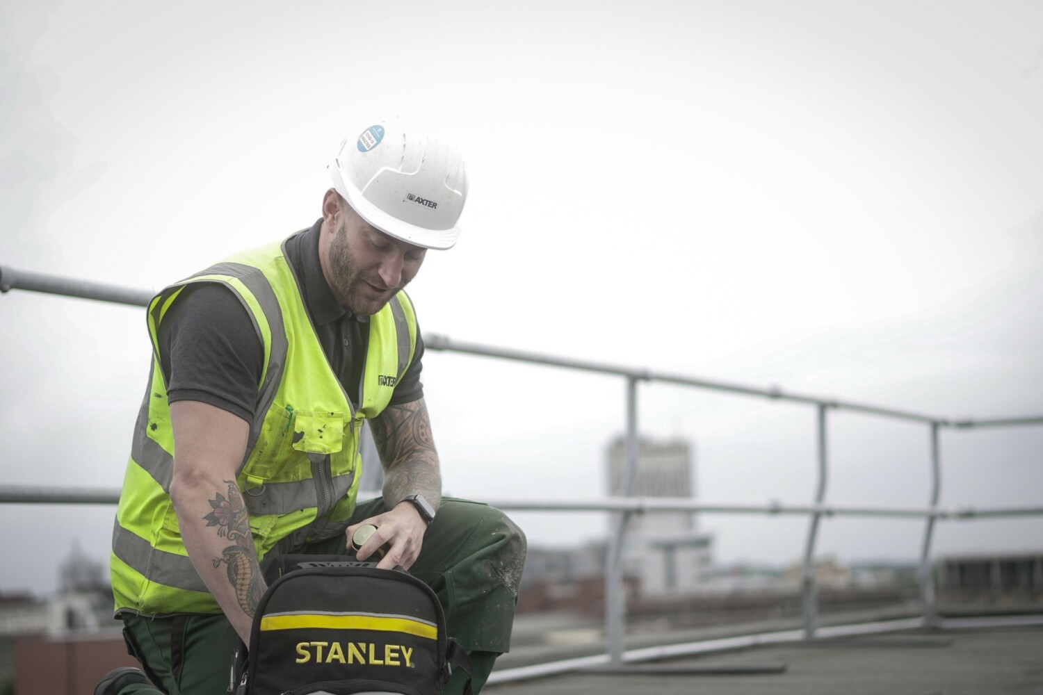 Construction worker in white hard hat and high-vis yellow vest kneeling beside green Stanley toolbox on rooftop with metal railings.