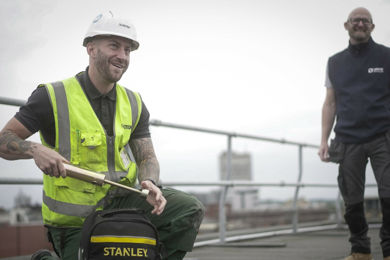 Construction worker in hi-vis jacket and hard hat kneeling beside green Stanley toolbox, with colleague in dark clothing standing behind metal railing.