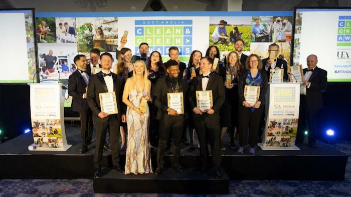 Large group of formally dressed people holding certificates on stage with blue lighting and presentation displays in background.