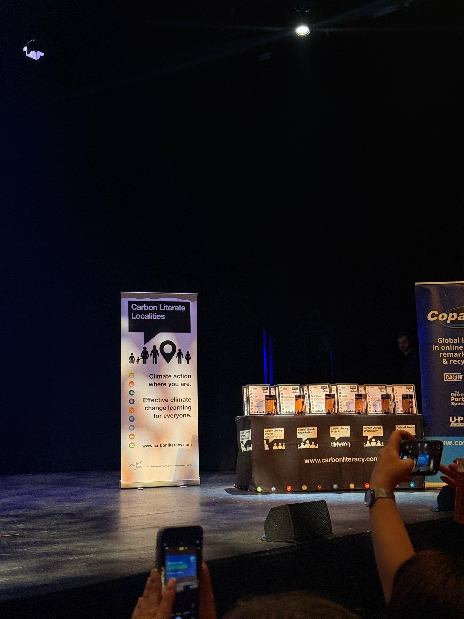 Conference stage with white banner reading "Carbon Literate Locations" and illuminated display booth in darkened venue.