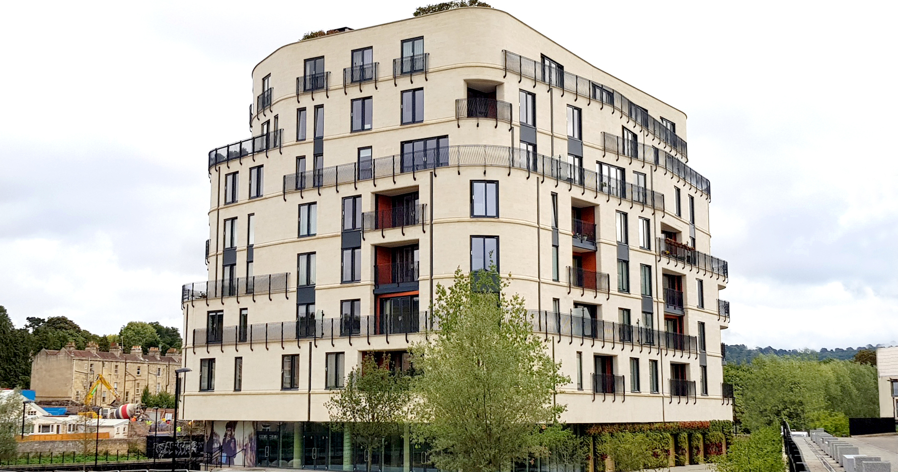 Modern curved apartment building with cream-coloured facade, dark window frames, and red balcony accents. Green tree in foreground, cloudy sky background.