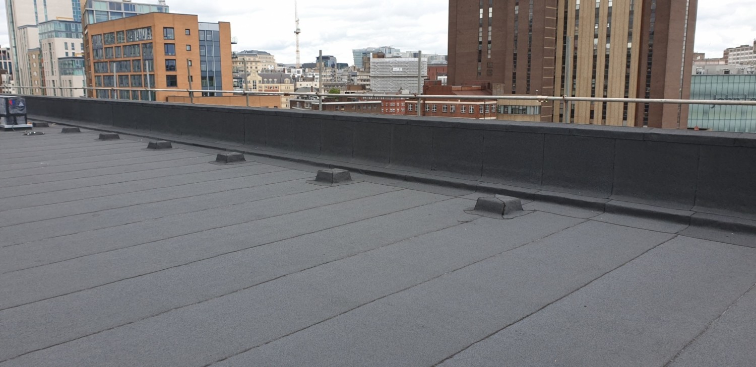 Grey flat roof with metal edging and drainage outlets, overlooking urban buildings including brick and modern structures.