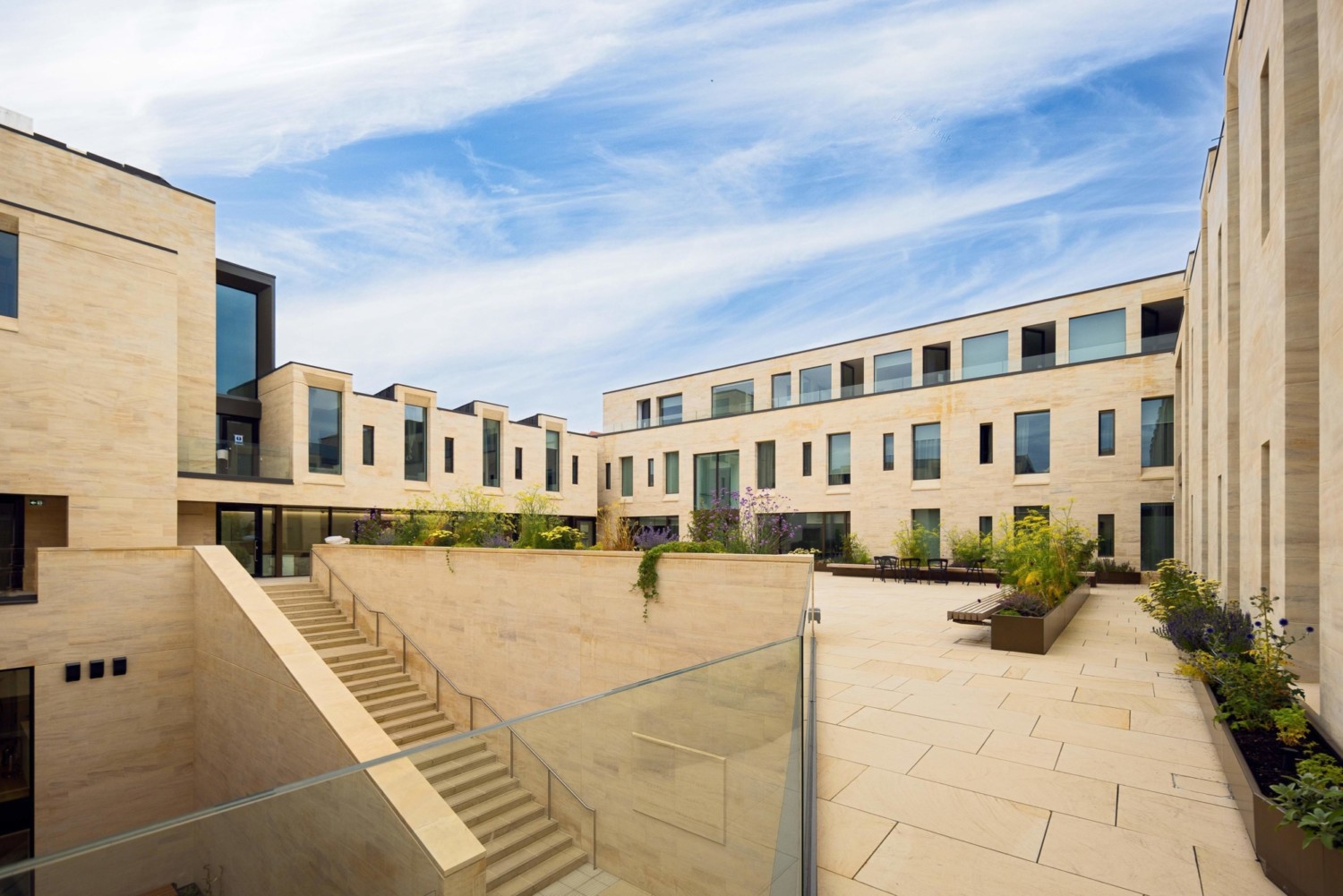 Modern residential courtyard with cream brick buildings, concrete steps, planted areas, and blue sky with white clouds overhead.