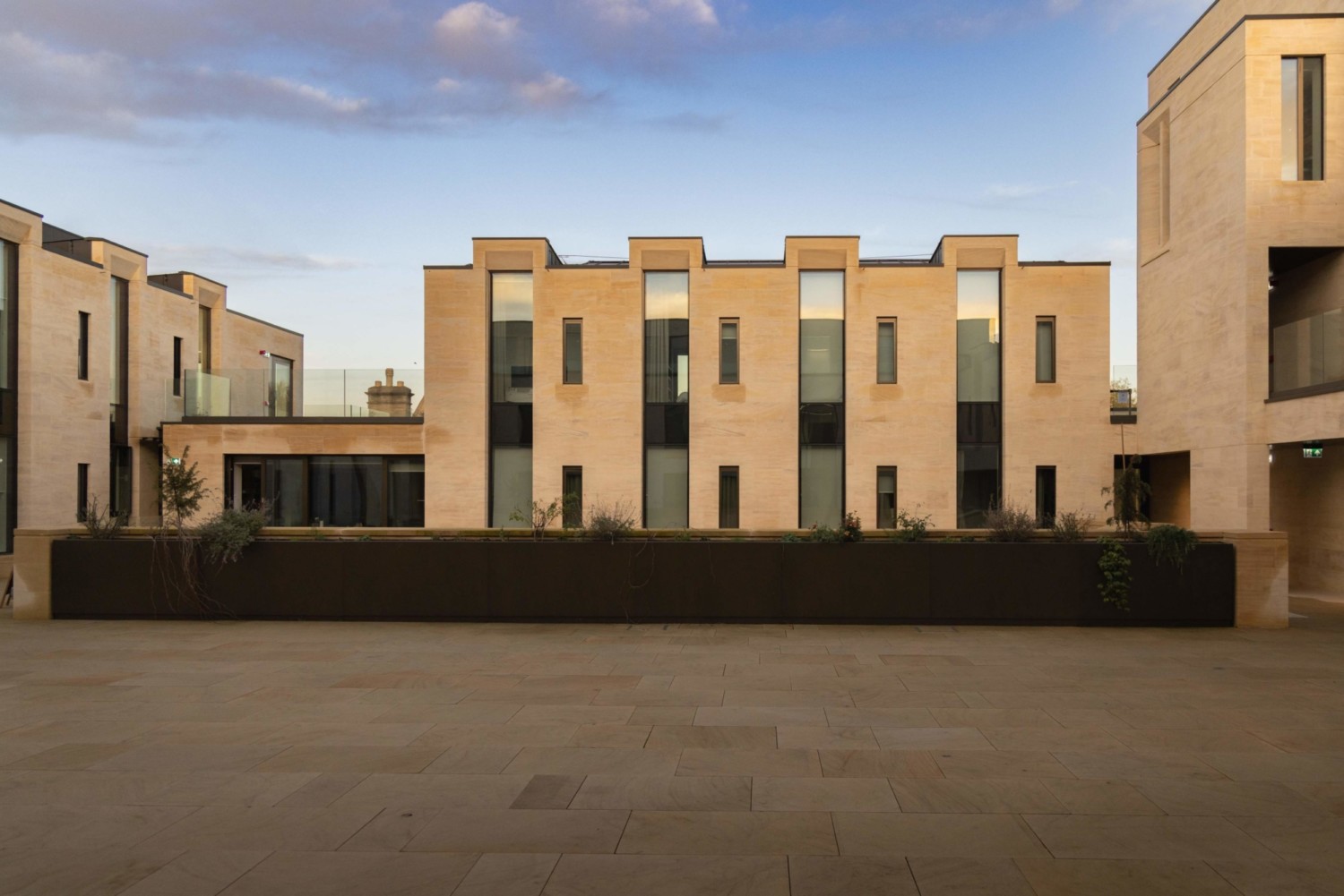 Modern residential development with beige brick facades, dark window frames, and crenellated rooflines under cloudy blue sky.
