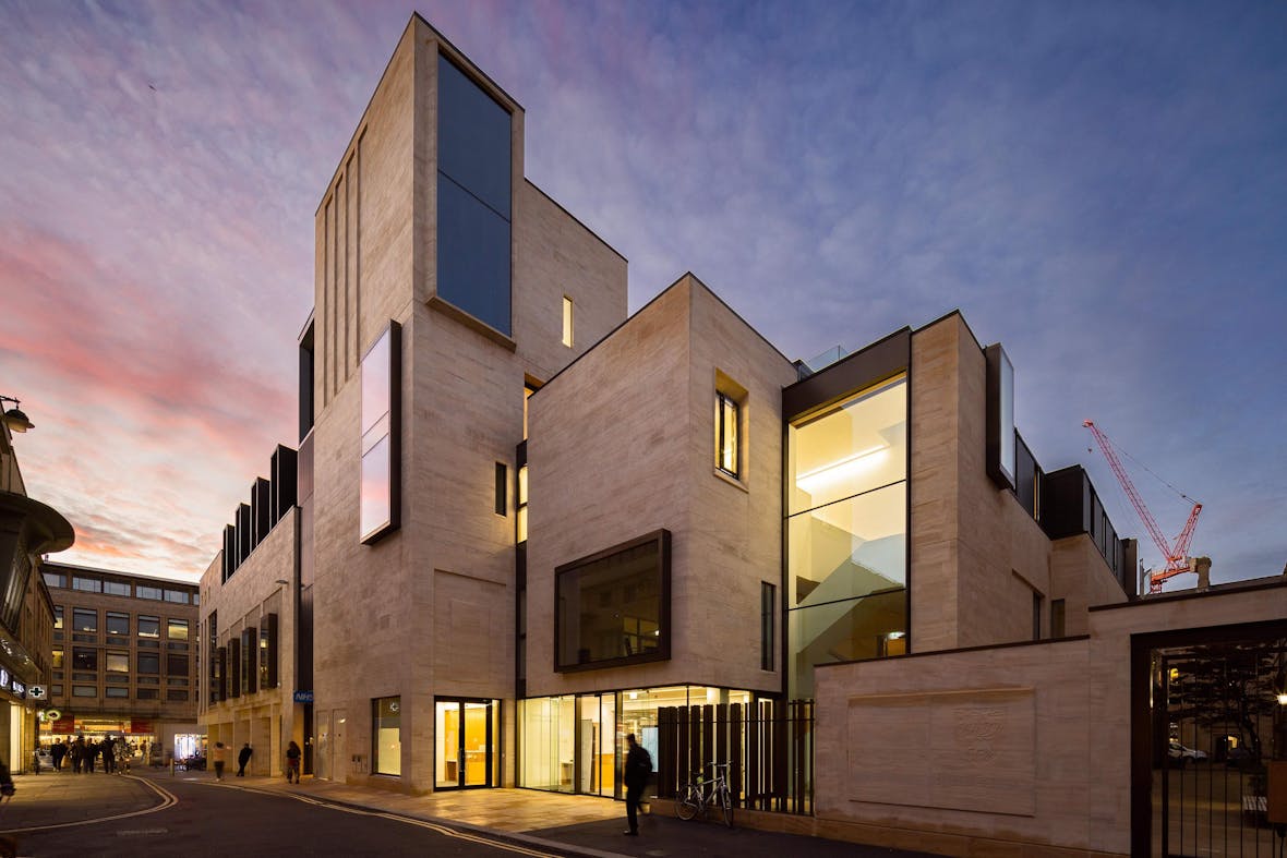 Modern concrete building with angular geometric forms, illuminated windows, and purple-pink twilight sky backdrop.