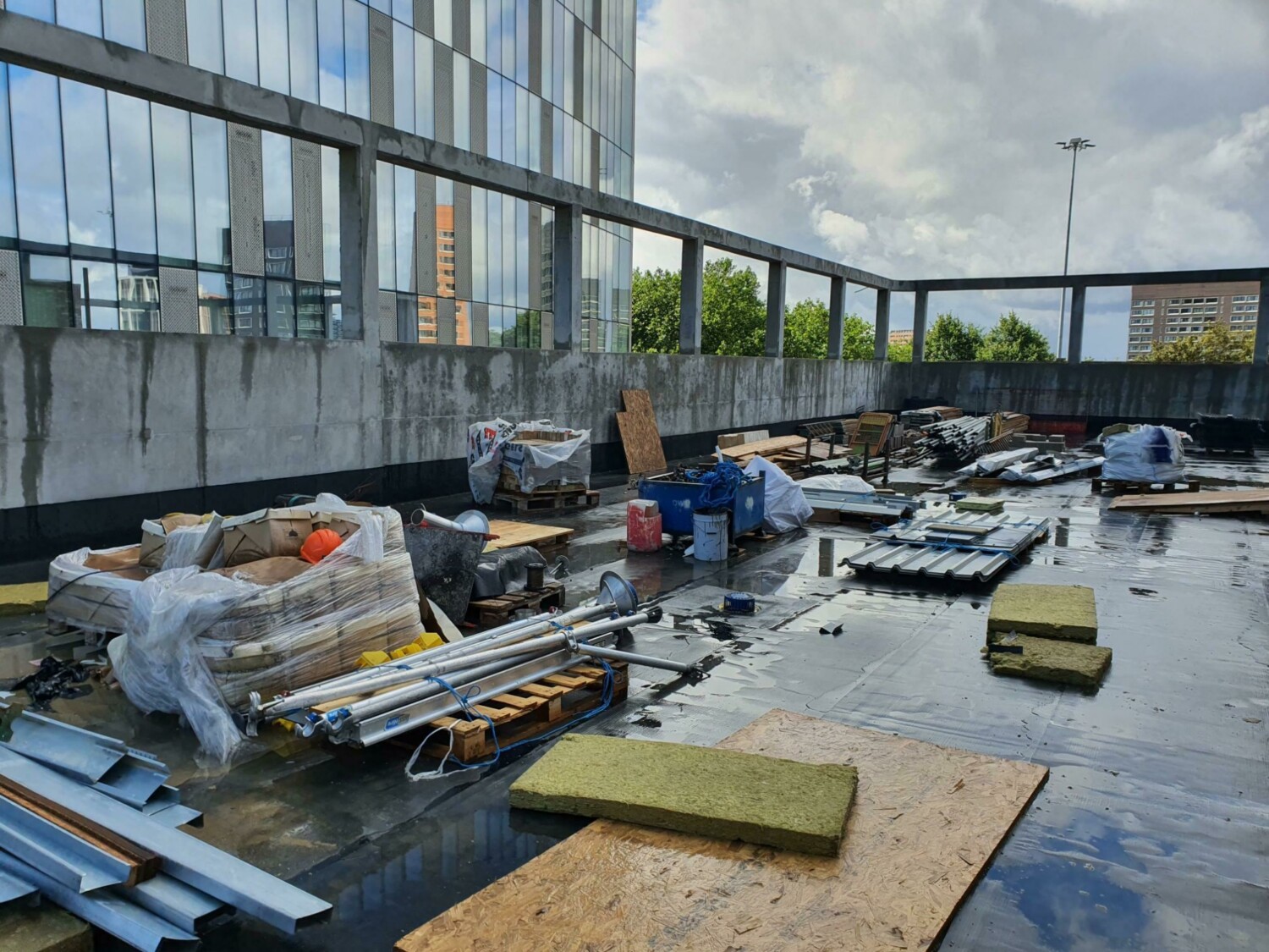 Construction materials and equipment scattered across wet rooftop with glass building facade and cloudy sky in background.
