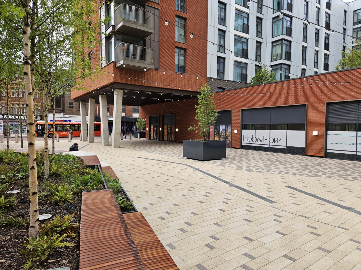 Modern residential development with red brick and white buildings, paved courtyard, wooden benches, planted areas, and ground-floor retail units.