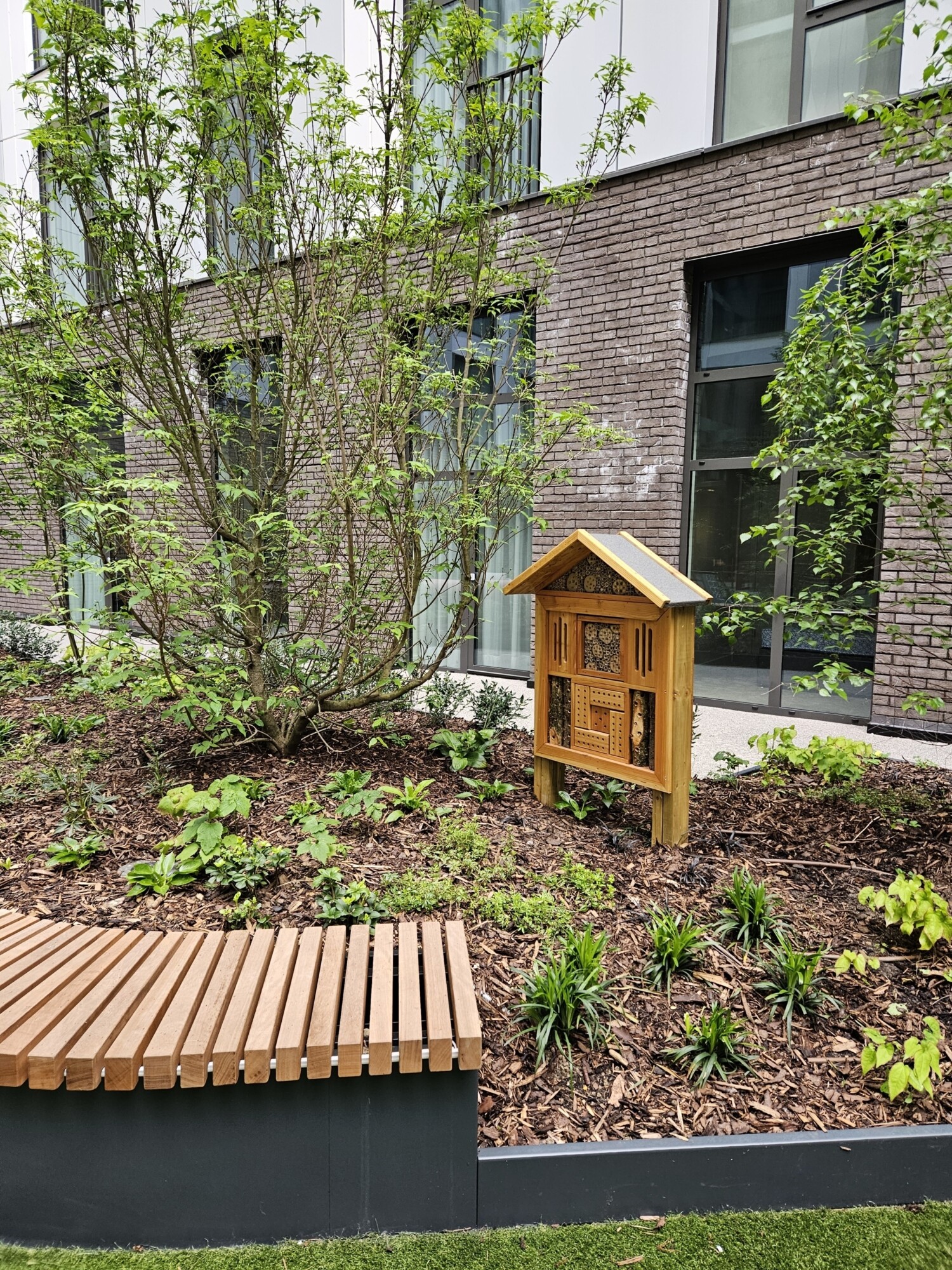 Modern brick building with wooden book exchange box in landscaped garden area, wooden bench and young trees visible.