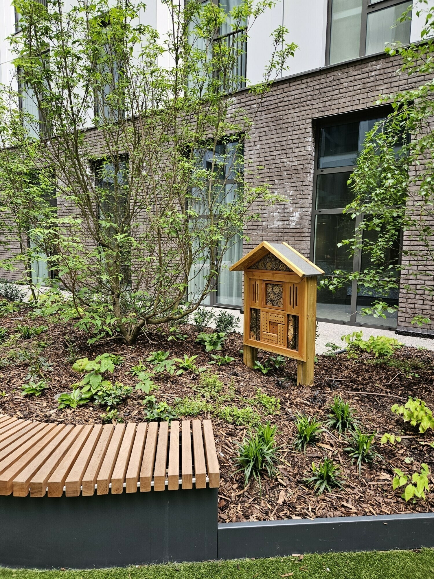 Modern brick building with large windows, wooden slatted bench, small wooden cabinet structure, and landscaped garden beds with green plants.