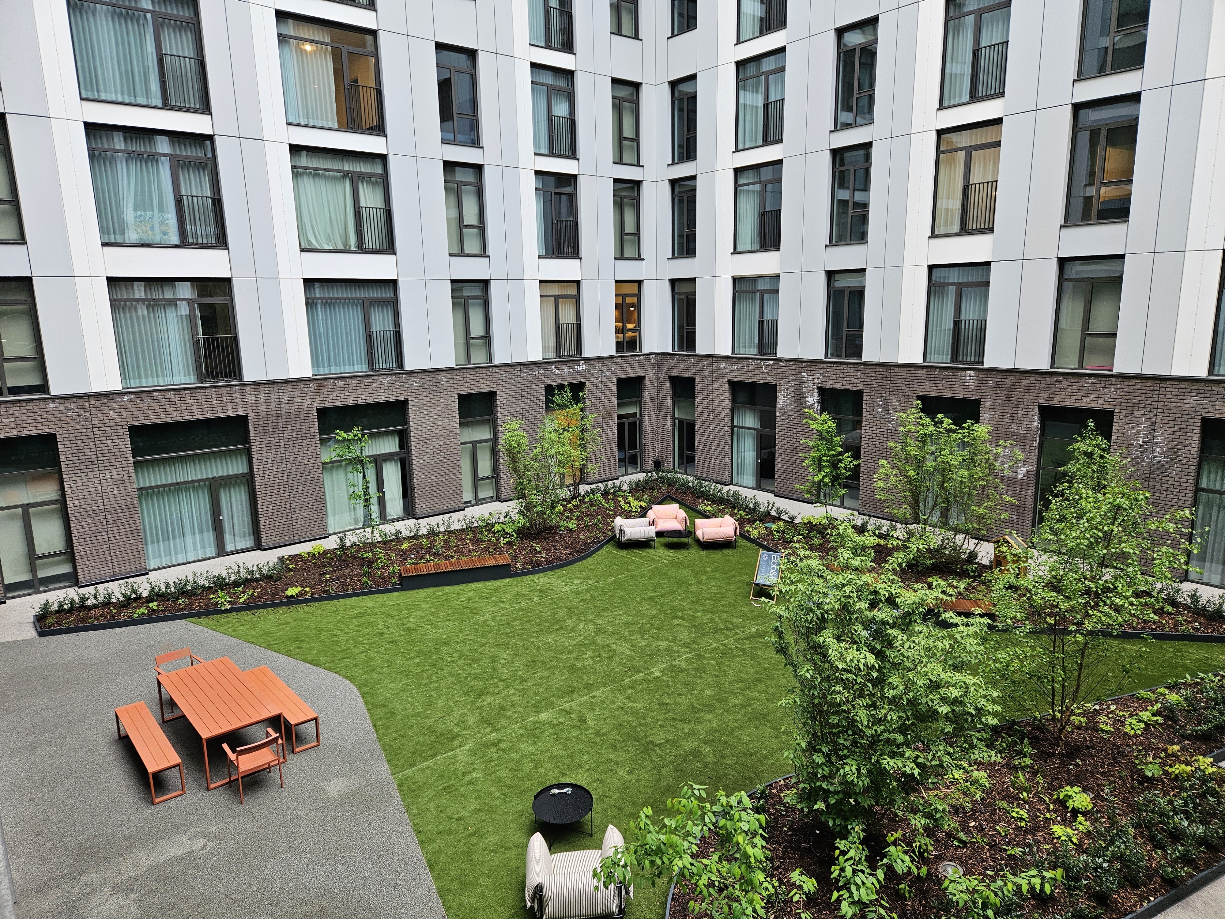 Modern apartment courtyard with white upper floors, brown lower level, central lawn, landscaped borders, orange outdoor furniture.