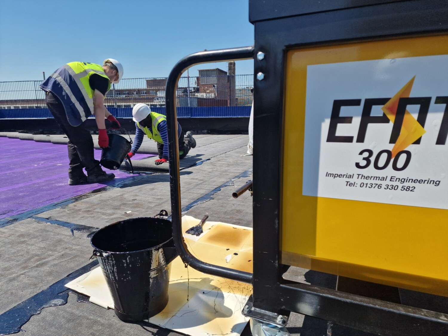 Construction workers in high-vis jackets laying purple roofing material on flat roof, with yellow EFT1 300 equipment in foreground.